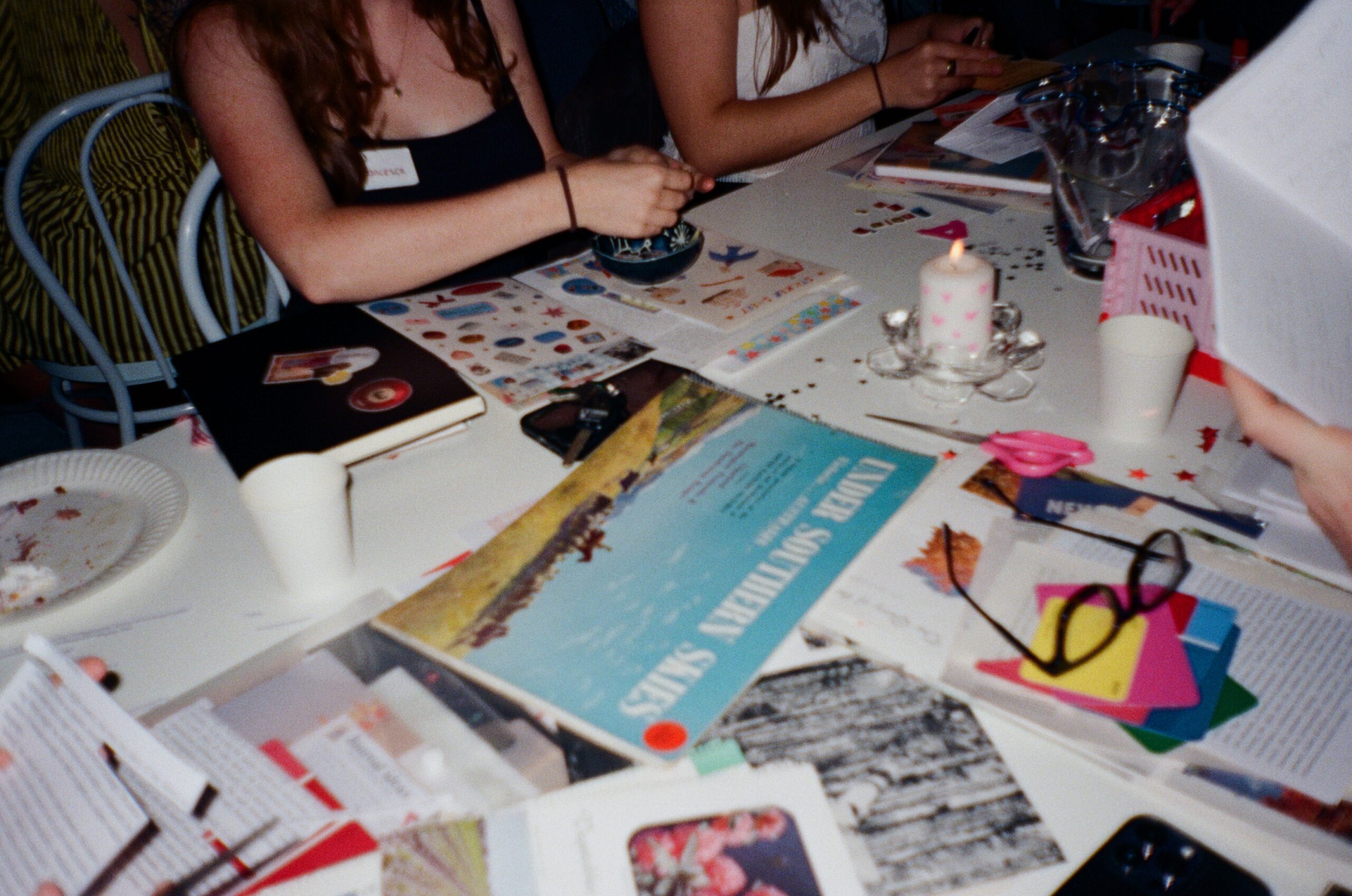 A table is scattering with old magazines and glue with people scrap booking.
