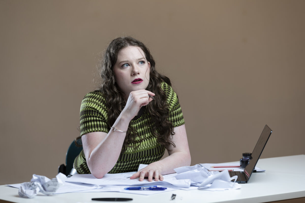 A young woman in a green and black shirt sits at a table strewn with papers.